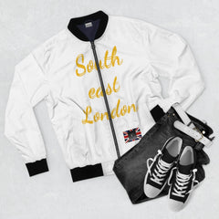 White bomber jacket with 'South east London' gold lettering, black collar, and UK flag patch, styled with dark jeans, white belt, and black sneakers.
