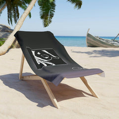 Black beach towel with white graphic design draped over a wooden lounge chair on sandy beach
